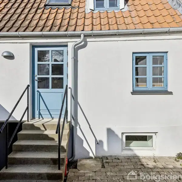 Husfacade med hvidmalet væg og blå dør samt vinduer. En trappe med sorte gelændere leder til døren. Omgivet af flisebelagt gårdsplads og charmerende, rødt tegltag.