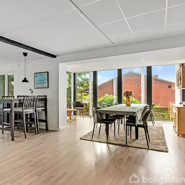 Spisebord med stole er placeret ved glasvæg, der giver udsigt til have og rødt murstenshus. Moderne åbent køkken- og spisestueområde med lys trægulv og minimalistisk indretning.