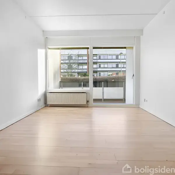 Et tomt værelse med lyse trægulve og hvide vægge. En radiator står under et stort vindue, der åbner ud mod en balkon med udsigt over en bygning udenfor.