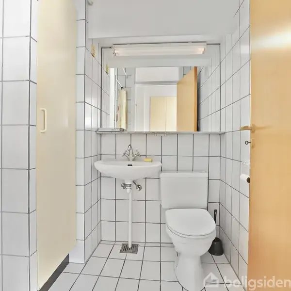 Et hvidt toilet og håndvask med spejl på flisebeklædt badeværelse. En bruser med glasdør er til venstre, mens der er en åben dør til højre.