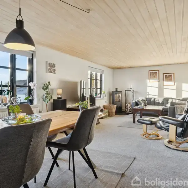 Spisestuebord med stole i et lyst rum; vinduer giver dagslys, og stuehjørne med sofa og pejs. Grønne planter dekorerer rummet, som har træloft og gulvtæppe.