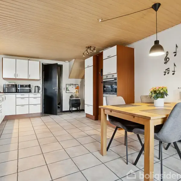 Køkken med hvide skabe og sort bordplade, indbygget ovn og mikroovn. Der er et spisebord med grå stole. En væg er dekoreret med musiknoter, blomster på bordet.