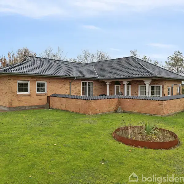 En enetages murstensvilla med mørkt tegltag står omgivet af en velplejet græsplæne med få træer og buske. Foran huset er der en hæk og en rund plantebed.