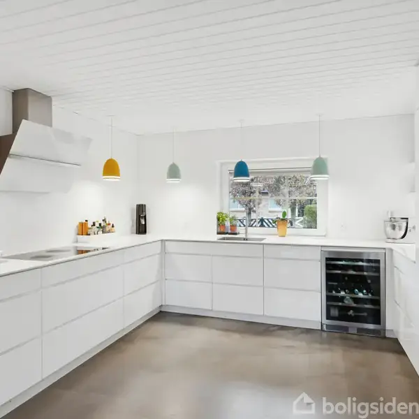 Hvidt køkken med moderne hvide skabe. Farverige pendel-lamper hænger over bordpladen. Et vindue med udsigt til haver fører lys ind. En vinreol og en ovn er indbygget.
