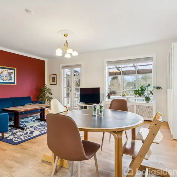 Spisebord med stole står i stue med trægulv. Sofa og tæppe ved rød væg. Vægmaleri og planter skaber en hyggelig atmosfære i den lyse stue med store vinduer.