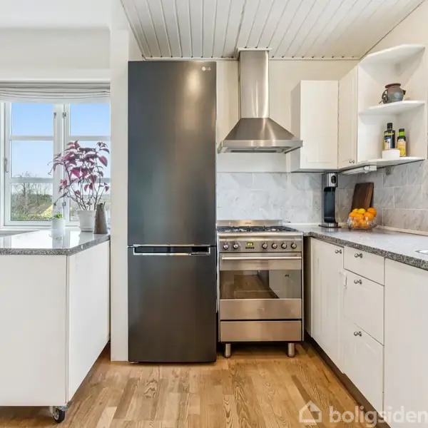 Et køkken med komfur og emhætte placeret mellem sorte køleskab og hvide skabe. Trægulve og hvide vægge kompletterer rummet. Vinduer til venstre tilfører naturligt lys.