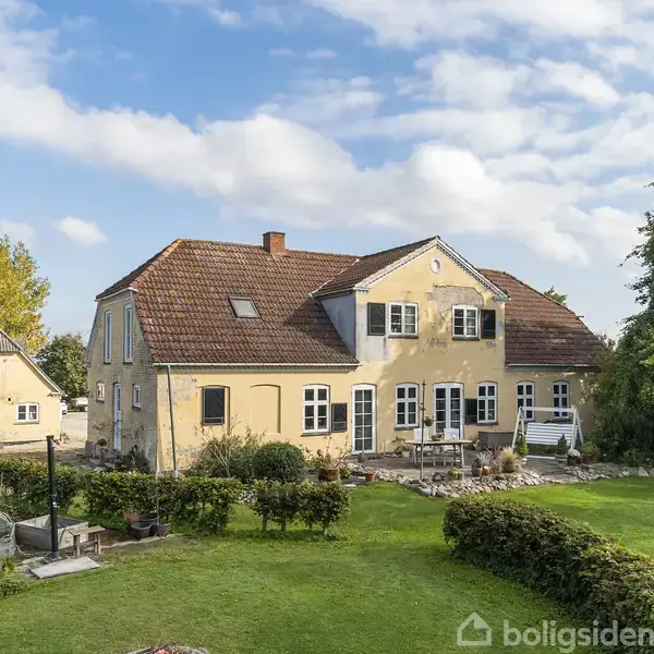 Et gult, to-etagers hus med en brun tegltag er omgivet af en velplejet have med græs, buske og træer. I baggrunden ses en mindre bygning.