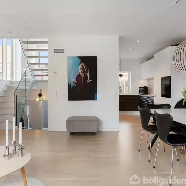 Stue med spisebord og moderne lamper; trappe til venstre. Lys træindretning med en sofa, vægkunst og planter skaber en luftig, minimalistisk atmosfære.