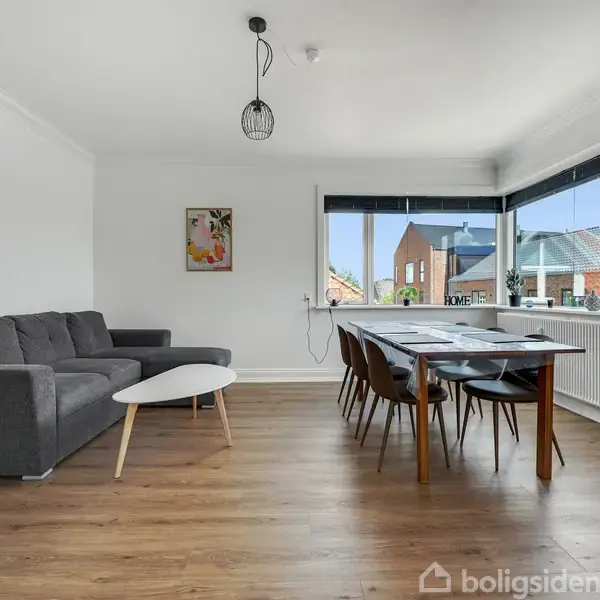 Grå sofa står i stuen ved siden af et hvidt sofabord. Et spisebord med stole står ved vinduet med udsigt til bygninger udenfor.