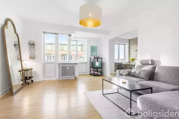 En lys stue med en grå sofa står ved et spisebord. En stor gulvspejl læner sig mod væggen. Et lille lampebord står ved et vindue.