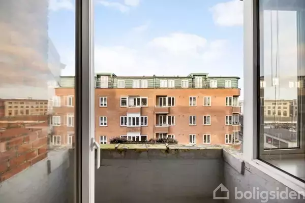 Et åbent vindue viser en altan med udsigt til en rød murstensbygning. Flere vinduer og altaner ses på bygningen under en blå himmel.