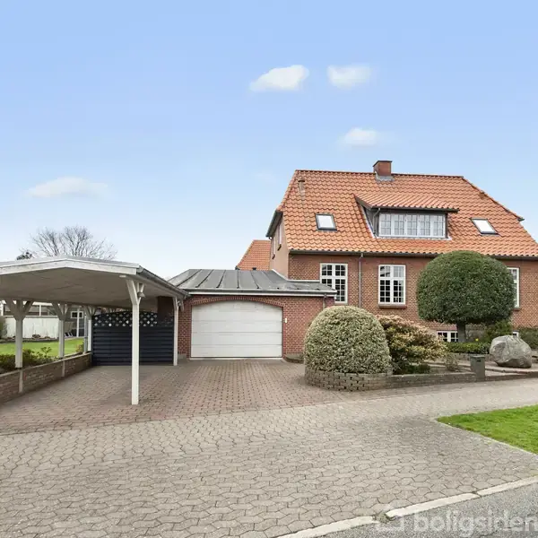 Rødt murstenshus med rødt tegltag står stille i et villakvarter. En carport og garage ligger til venstre, omkranset af havebede og buske. Klar himmel i baggrunden.