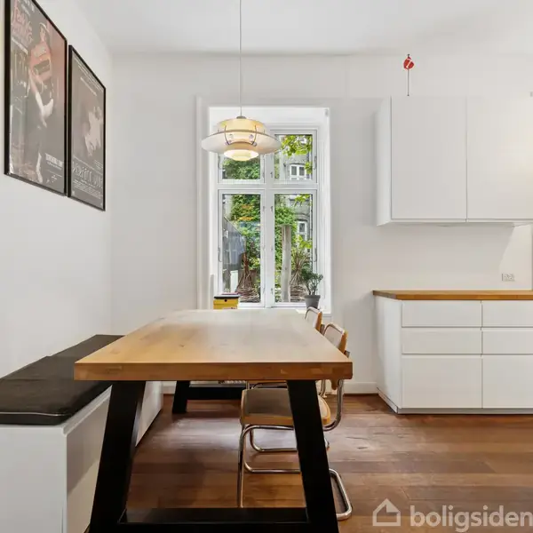 Et lyst køkken med et træbord og fire stole opstillet. På væggen hænger tre plakater. Til højre ses hvide skabe og et vindue med udsigt til haven.