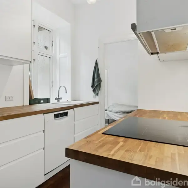 Et moderne køkken med hvide skabe og træbordplader. En indbygget kogeplade ses på øen, mens en ovn og emhætte er ovenover. Håndklæde hænger ved vasken i hjørnet.