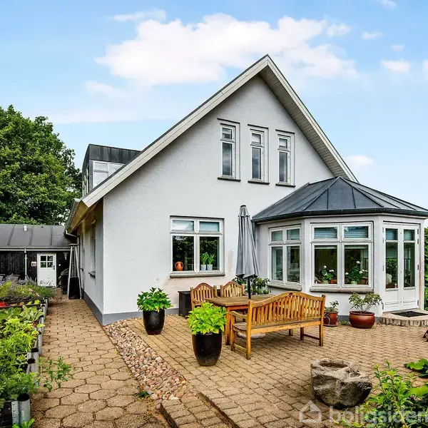 Hvidt hus med spidst tag står stille i en have med flisebelagt terrasse, hvor der er placeret en træbænk, krukker med planter og grønne buske omkring.