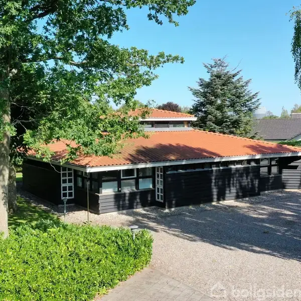 Hus med mørkt træværk og rødt tag står stille omgivet af træer og grusadgang i en solrig have.