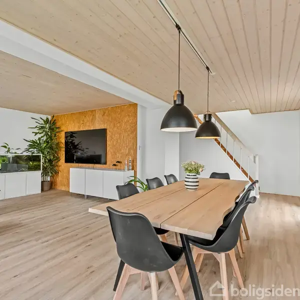 Et spisebord med sorte stole står under tre pendellamper i en lys, moderne stue med træloft. Til venstre ses en blå sofa, akvarium og planter ved en TV-skærm.