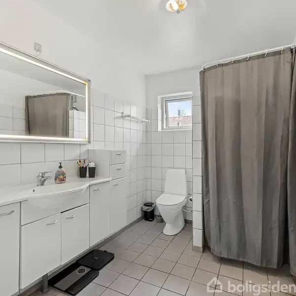Badeværelse med hvidt skabsarrangement, håndvask og spejl til venstre. Bruser med gråt gardin i hjørnet. Toilet ved siden af lille vindue. Lyse fliser dækker gulv og vægge.