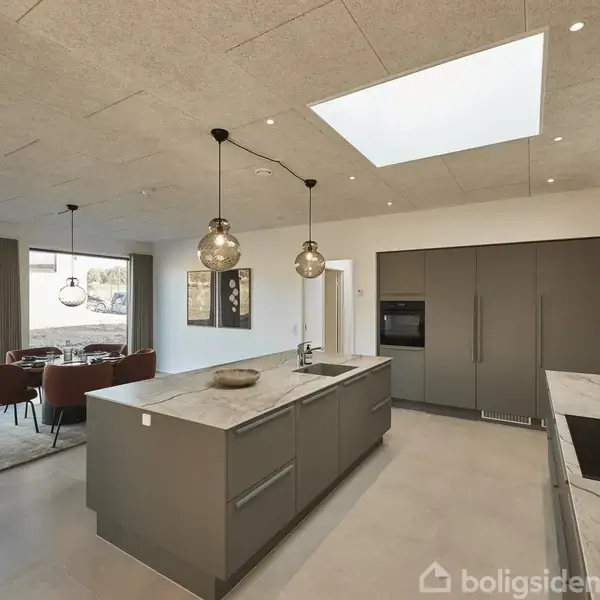 Køkken med en central ø, dekorative lamper hængende over, fliser på gulvet, og vinduer i baggrunden giver udsigt til haven. Spiseområde med bord og stole placeret ved siden af.