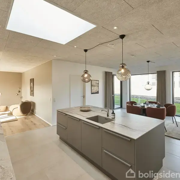 Køkkenø i moderne køkken med vask og bordplade, oplyst af to hængelamper; i baggrunden ses spiseplads med runde borde og stole foran store vinduer mod have.