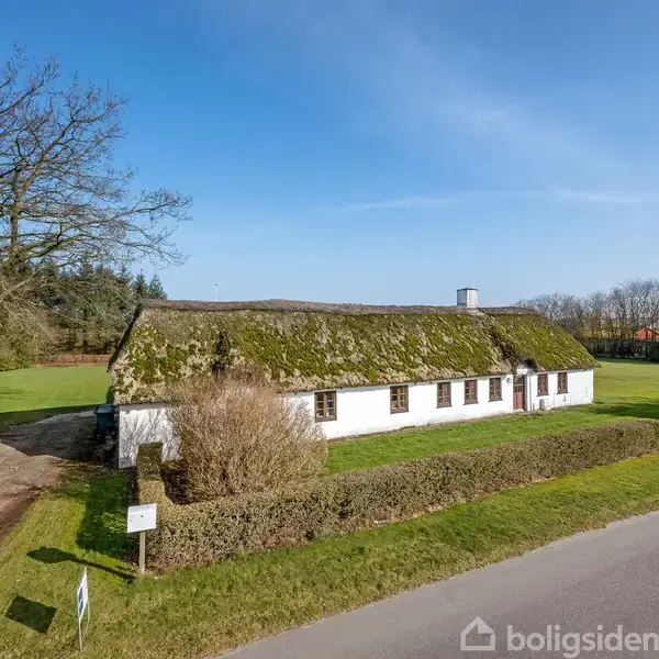 Et langstrakt, stråtækt hus står stille i et grønt, åbent landskab. Der er en stor træ ved siden af grusstien og velholdte hække omgiver ejendommen.