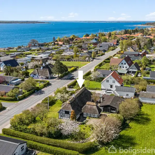 Bindingsværkshus med stråtag indikeret af pil, omgivet af grønne haver og andre boliger. Baggrund med udsigt over vandet langs kystlinjen og en vej, der skærer gennem landsbyen.