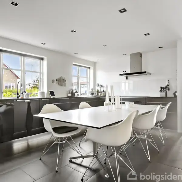Et moderne køkken med hvid bordplade og seks hvide stole står i et lyst rum. Store vinduer giver udsigt til en villa udenfor, og rum med grå fliser.