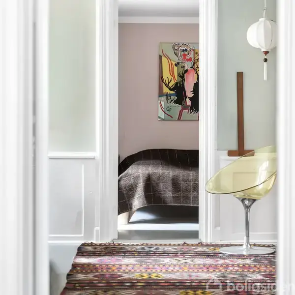 En seng dækket med et mørkligt tæppe står i et værelse. Et farverigt maleri hænger over sengen, mens en gennemsigtig stol er placeret i forgrunden.