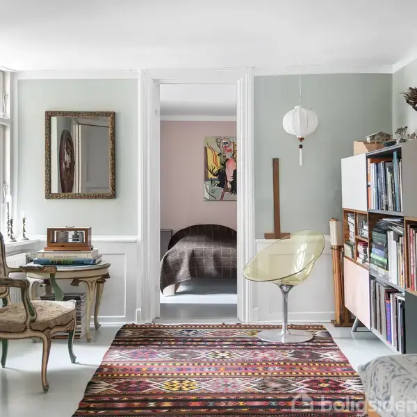 En stue med væg-til-væg-tæppe i farverigt mønster, en stol og et lille bord ved vinduet. Reol med bøger og lamper langs væggen. Åben dør til soveværelse.