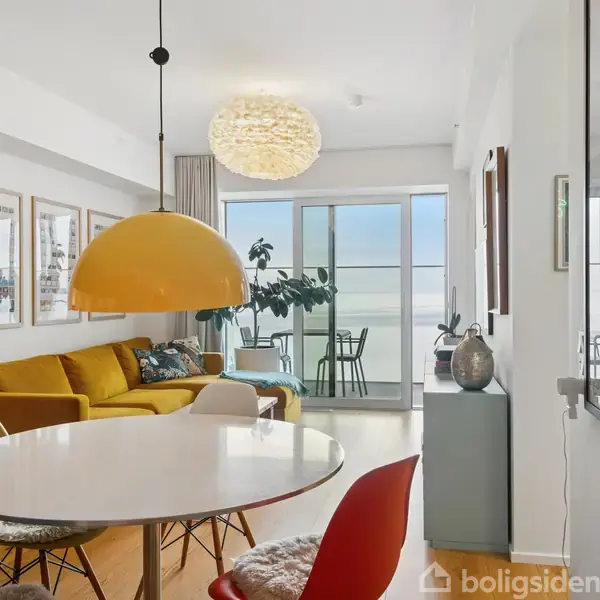 Et moderne stueområde med gul sofa og runde bord omgivet af farverige stole. Stor gul lampe hænger over bordet. I baggrunden ses glasdøre til en balkon.