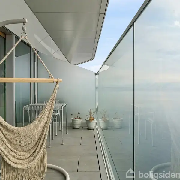 Hængekøjestol hænger på moderne altan med glaspaneler. I baggrunden står to stole ved et lille bord, og flere plantepotter, med udsigt over havet.
