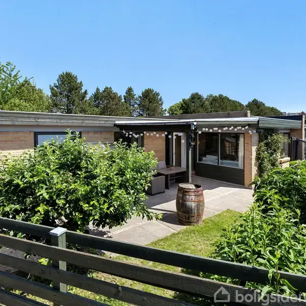 Et murstenshus med store vinduer står i en solrig have. Foran huset er en terrasse med bænkesæt og et stort træfad, omkranset af frodig vegetation og et plankeværk.