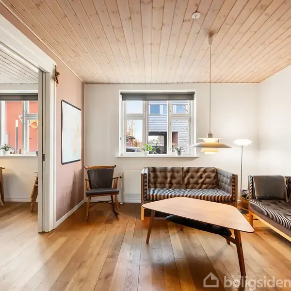 Stue med to sofaer i trærammer, sofabord og gulvlampe. Trægulv og loft. Store vinduer giver udsigt til gadens bygninger, mens et tilstødende rum ses gennem en døråbning til venstre.