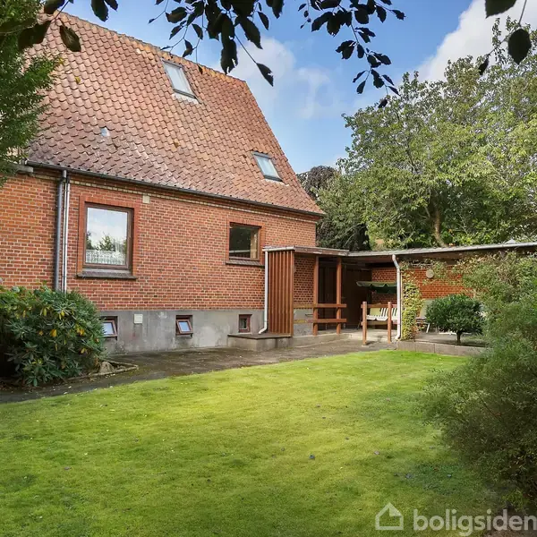 Et rødt murstenshus står stille omgivet af en velplejet græsplæne. Træer og buske omkranser bygningen, som har et skråt tegltag og flere vinduer.