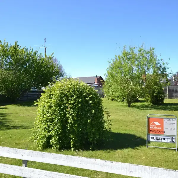 Græsplæne med en stor busk; ejendomsmæglerens skilt viser "EDC Jørgen Danielsen, Ejendomsmægler & valuar MDE, TIL SALG". Flere træer og huse i baggrunden, solrig dag.