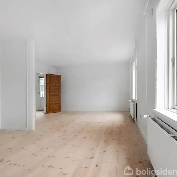 Et rummeligt, lyst rum med trægulv, hvor en trædør står åben og lader lys komme ind fra store vinduer på højre side. Ingen synlig tekst.