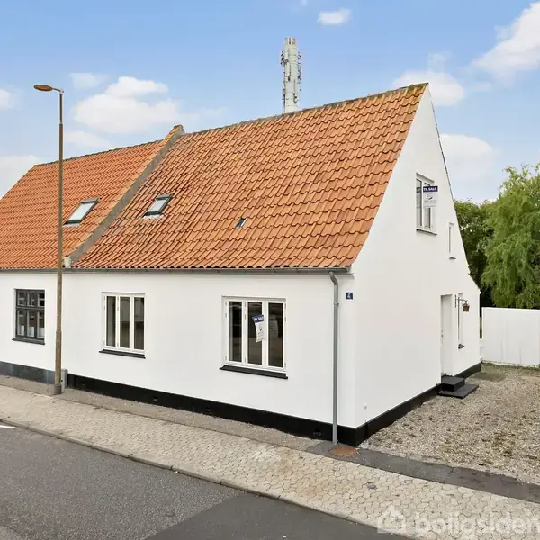 Hvidt hus med rødt tegltag står stille på fortovet ved vejkanten, omgivet af et lille grusareal og grøn vegetation i baggrunden.