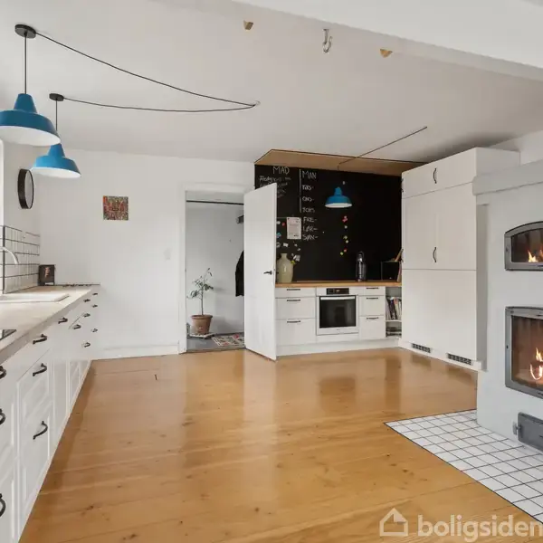 Pejseovn brænder lystigt i køkkenområdet, omgivet af hvide skabe og en træbordplade. Blå lamper hænger ned over rummet, der har flisebelagt gulv nær pejsen og trægulv ellers.