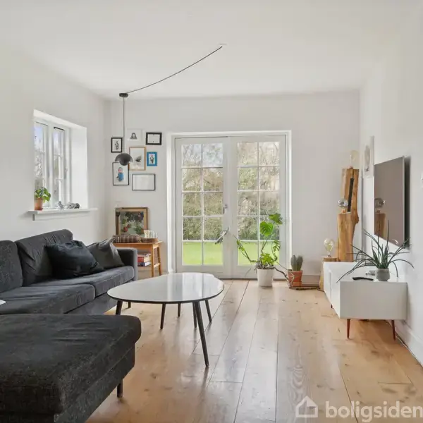 Sort sofa står i en lys stue med hvide vægge, træbord, og billeder på væggen. Store glasdøre fører ud til en have, og planter tilføjer grønne detaljer.