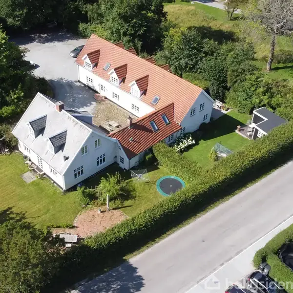 To huse med røde tage ligger i et grønt parcelhuskvarter. En trampolin er placeret i haven, omgivet af hække og træer. En vej løber langs forhaven.