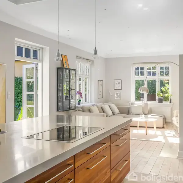 Køkkenø med vask og kogeplade står centralt i et lyst rum, åbent mod en stue med sofa og store vinduer, der giver udsigt til en terrasse og grønne omgivelser.