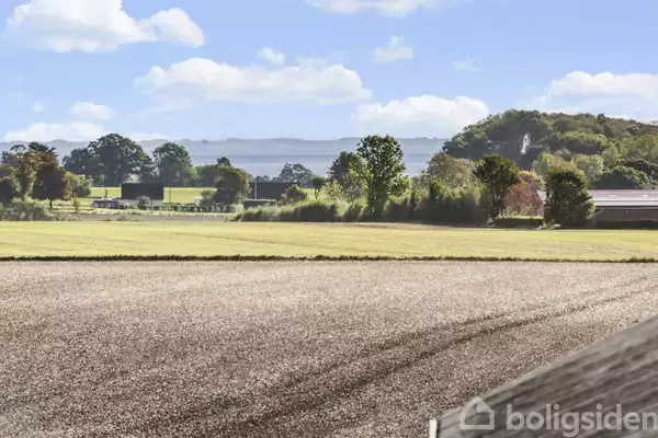 En mark med tydelige traktorspor breder sig i forgrunden. I baggrunden ses grønne marker og træer under en blå himmel med lette skyer.