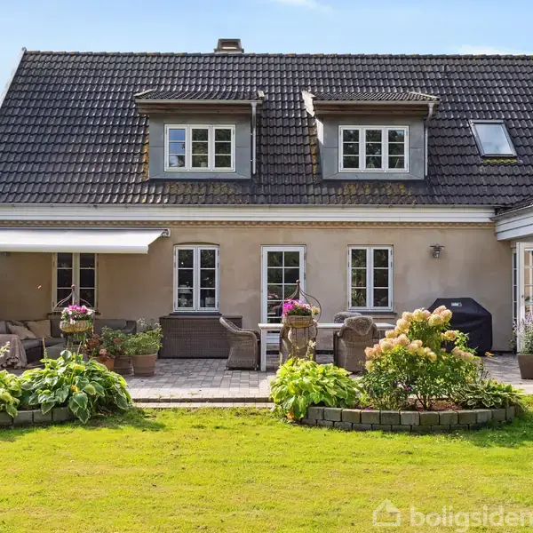 Et hus med mørkt tegltag har en stor terrasse med havemøbler og blomsterkrukker. Terrassen er omgivet af en velplejet græsplæne og frodig hæk til venstre.