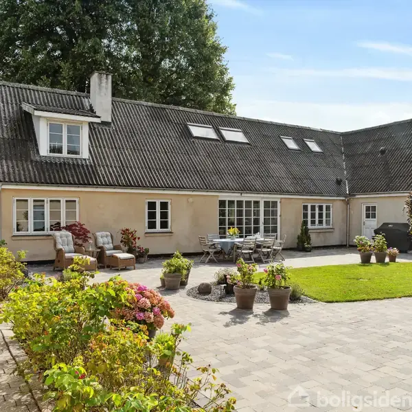 Hus med skråt tag omgivet af blomsterpotter og havemøbler på en flisebelagt terrasse. Omkring er grønne planter og buske med en stor træ i baggrunden.