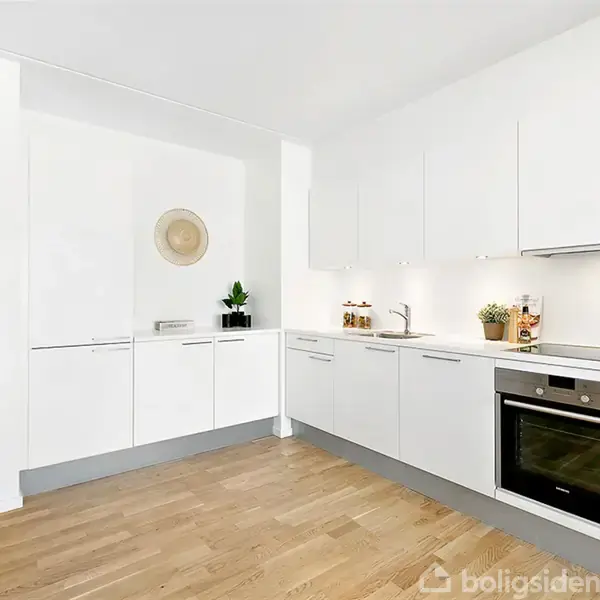 Et moderne køkken med hvide skabe og trægulv. Der er en indbygget ovn, køkkenredskaber på bordpladen samt en plante og en dekorativ kurv på væggen. En åbning fører til en lys stue.