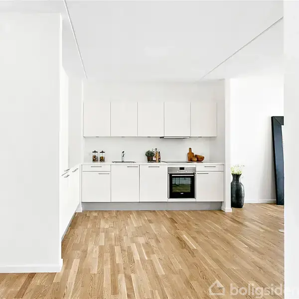 Et moderne køkken med hvide skabe og indbygget ovn står i en lys lejlighed med trægulve. Til venstre ses en åben dør, der fører til et soveværelse.