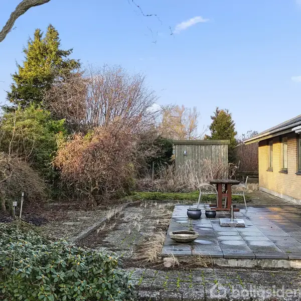 Hus med en flisebelagt terrasse omgivet af buske og træer i en have. Et bord og krukker står på terrassen. En træbeklædt skur ses i baggrunden under en klar blå himmel.