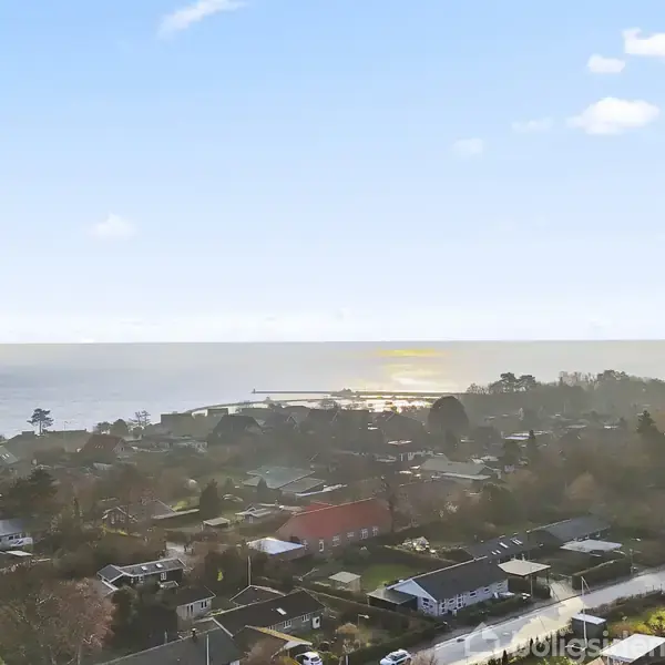 Kystby omgivet af huse med haver, beliggende ved en bred kystlinje. Sollys reflekterer på havets overflade, mens himlen er delvist dækket af skyer.