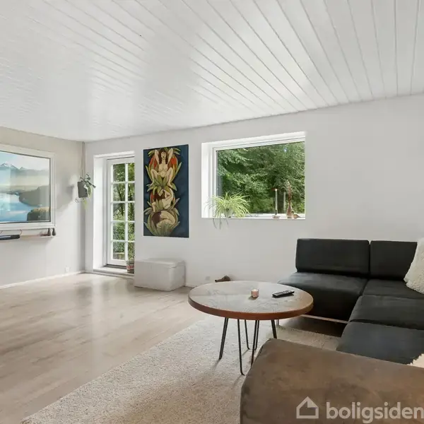 En lys stue med sort hjørnesofa og rundt træbord står på et lyst gulvtæppe. Et TV viser et landskab. Væggen har kunstværker og store vinduer med udsigt til natur.