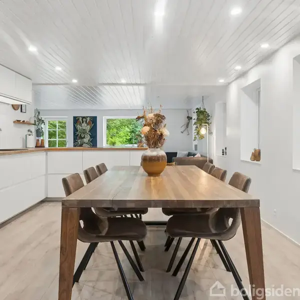 Et træspisebord med otte stole står i et moderne køkkenalrum. Rummet er lyst med hvide vægge og lofter, store vinduer og planter på borde og hylder, hvilket skaber en indbydende atmosfære.